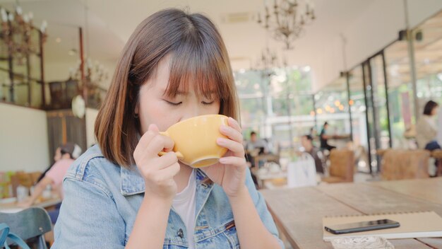 Mujer asiática independiente del negocio que bebe la taza caliente de té verde o de café mientras que se sienta en la tabla en café.