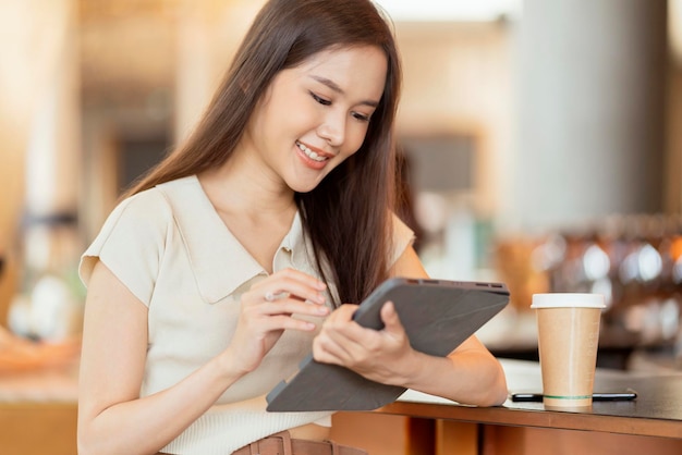 Mujer asiática independiente nómada digital mano usando una tableta que trabaja de forma remota en cualquier lugar de trabajo sentada en el mostrador cerca de la barra de café para hacer contacto con el cliente en el extranjero con un acuerdo de éxito pacífico sonriente alegre