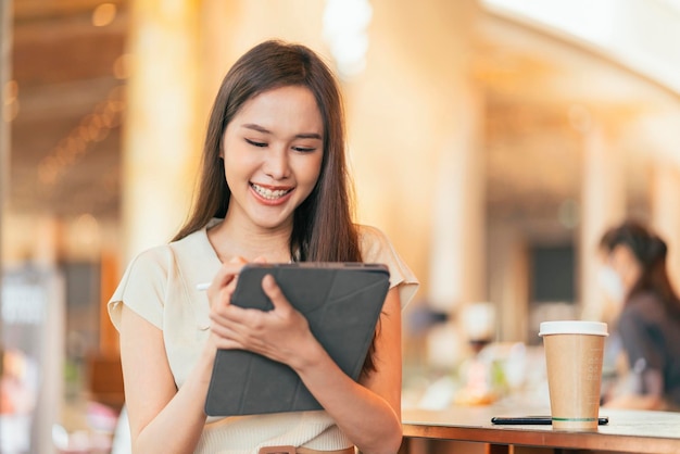 Mujer asiática independiente nómada digital mano usando una tableta que trabaja de forma remota en cualquier lugar de trabajo sentada en el mostrador cerca de la barra de café para hacer contacto con el cliente en el extranjero con un acuerdo de éxito pacífico sonriente alegre