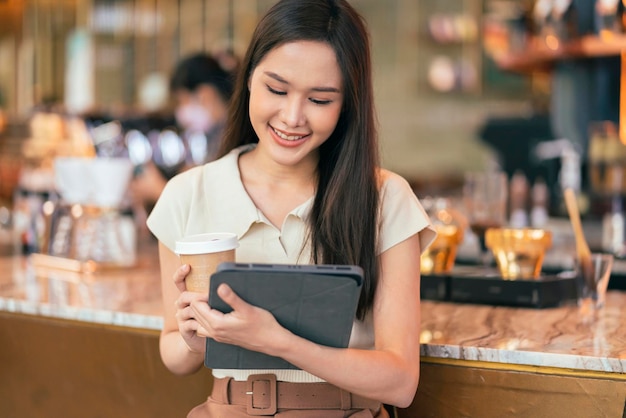 Mujer asiática independiente nómada digital mano usando una tableta que trabaja de forma remota en cualquier lugar de trabajo sentada en el mostrador cerca de la barra de café para hacer contacto con el cliente en el extranjero con un acuerdo de éxito pacífico sonriente alegre