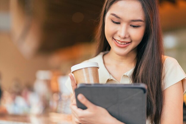 Mujer asiática independiente nómada digital mano usando una tableta que trabaja de forma remota en cualquier lugar de trabajo sentada en el mostrador cerca de la barra de café para hacer contacto con el cliente en el extranjero con un acuerdo de éxito pacífico sonriente alegre