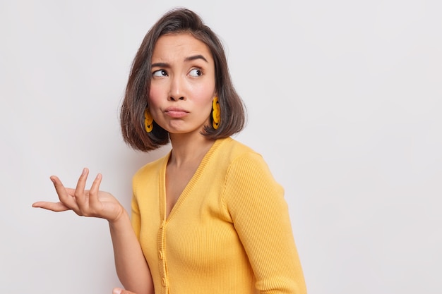 La mujer asiática indignada y disgustada concentrada tiene una expresión triste que levanta la mano con una mirada desorientada, usa aretes de puente amarillo posa contra la pared blanca, copia el espacio para texto o promoción