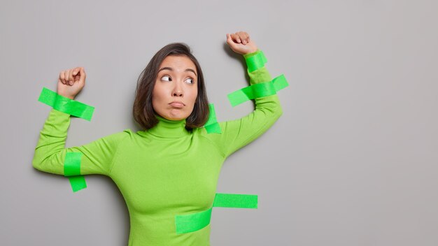 La mujer asiática infeliz frustrada en el cuello alto verde plastó a la pared gris mantiene los brazos levantados tiene la expresión de la cara de descontento siendo atrapada aislada sobre la pared gris