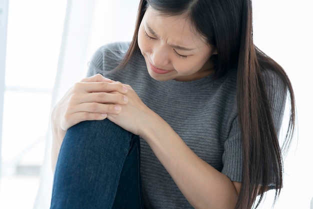 Una mujer asiática infeliz toca la rodilla de la pierna con dolor y lesiona el fondo blanco