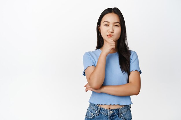 Mujer asiática intrigada que parece pensativa pensando en algo que tiene una suposición de pie en una camiseta azul sobre fondo blanco