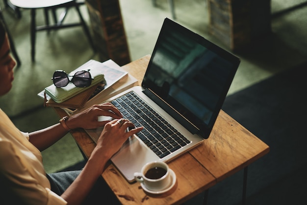 Mujer asiática irreconocible sentada en la cafetería y trabajando en la computadora portátil