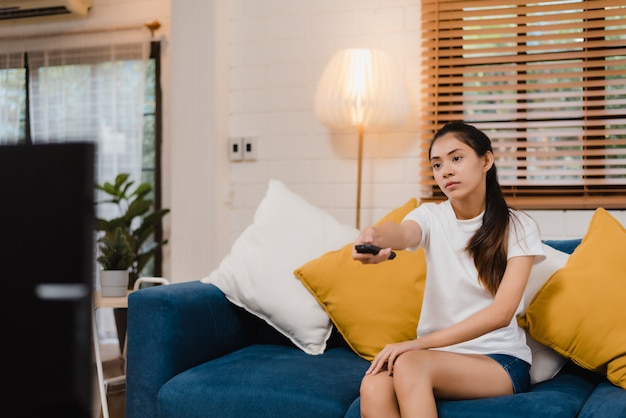 Mujer asiática joven del adolescente que ve la TV en casa, mentira feliz de la sensación femenina en el sofá en sala de estar.