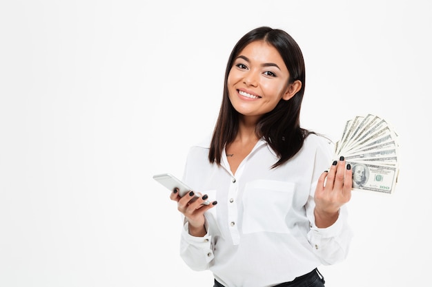Mujer asiática joven alegre que sostiene el dinero usando el teléfono móvil.