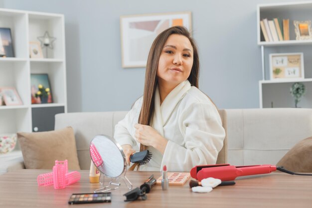 Mujer asiática joven en bata de baño con cabello largo oscuro sentada en el tocador en casa haciendo rutina de maquillaje matutino cepillándose el cabello con un cepillo para el cabello sonriendo