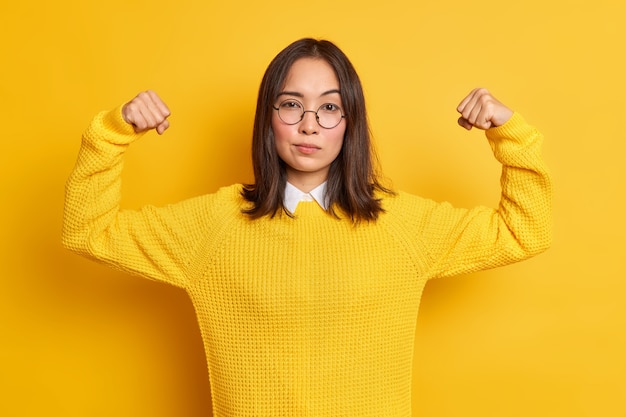 La mujer asiática joven confiada muestra los músculos del brazo se siente como un héroe demuestra su poder y fuerza se ve seriamente viste un suéter de gafas ópticas redondas.