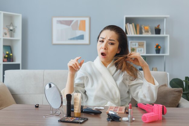 Mujer asiática joven confundida con toalla en bata de baño sentada en el tocador en el interior de la casa tocando su cabello disgustado haciendo rutina de maquillaje matutino