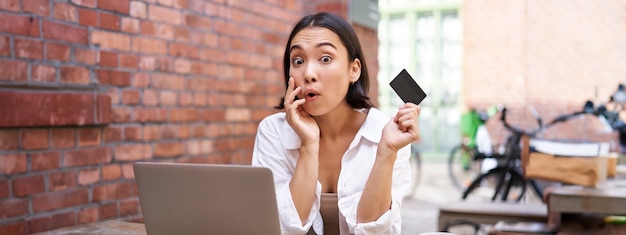 Mujer asiática joven y elegante haciendo compras en línea sentada con tarjeta de crédito y computadora portátil comprando