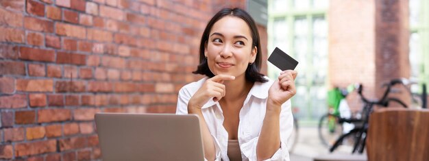 Mujer asiática joven y elegante haciendo compras en línea sentada con tarjeta de crédito y computadora portátil comprando