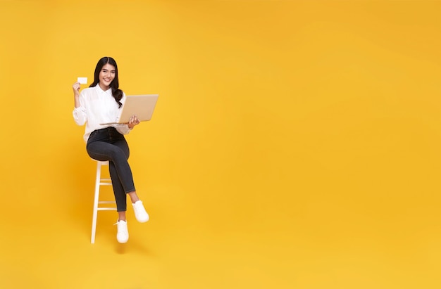 Mujer asiática joven feliz mostrando tarjeta de crédito y usando compras en línea de computadora