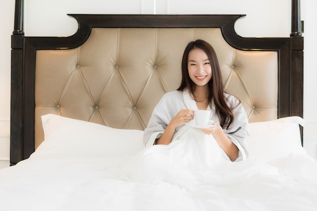 La mujer asiática joven hermosa del retrato despierta con sonrisa feliz y la taza de café en cama en el dormitorio inter