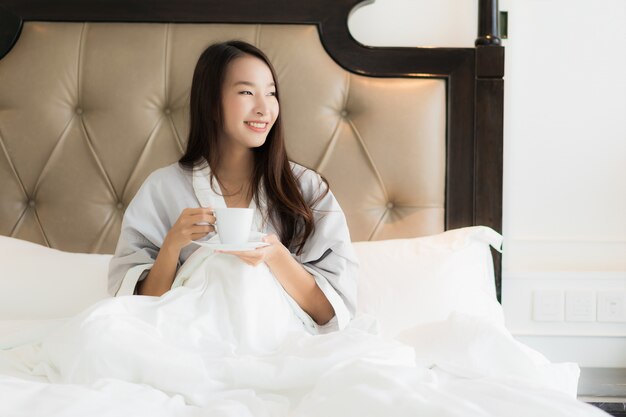 La mujer asiática joven hermosa del retrato despierta con sonrisa feliz y la taza de café en cama en el dormitorio inter