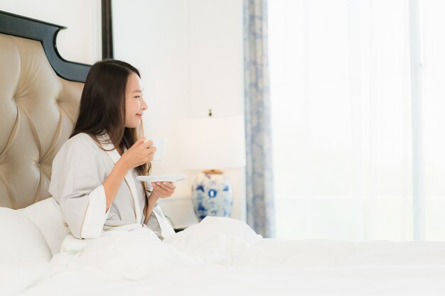 La mujer asiática joven hermosa del retrato despierta con sonrisa feliz y la taza de café en cama en el dormitorio inter