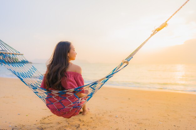 Mujer asiática joven hermosa del retrato que se sienta en la hamaca con el mar y el oce cercano felices de la playa de la sonrisa