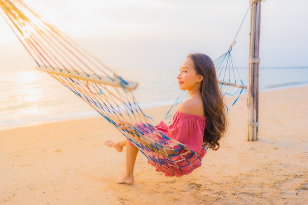 Mujer asiática joven hermosa del retrato que se sienta en la hamaca con el mar y el oce cercano felices de la playa de la sonrisa