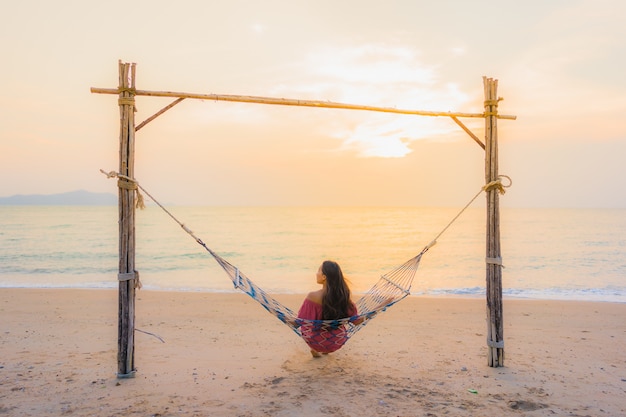 Mujer asiática joven hermosa del retrato que se sienta en la hamaca con el mar y el oce cercano felices de la playa de la sonrisa