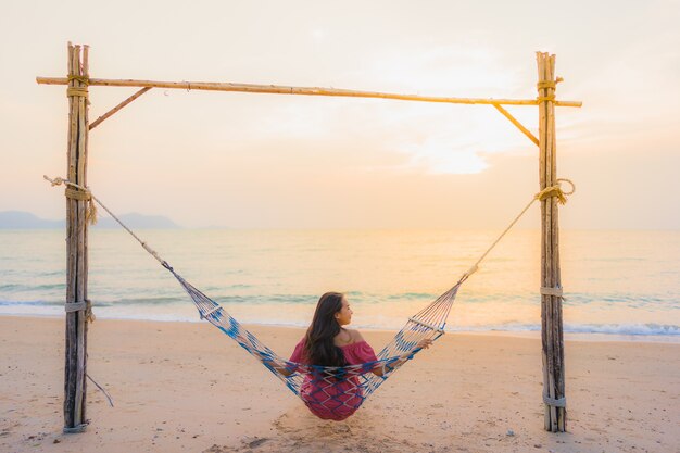 Mujer asiática joven hermosa del retrato que se sienta en la hamaca con el mar y el oce cercano felices de la playa de la sonrisa