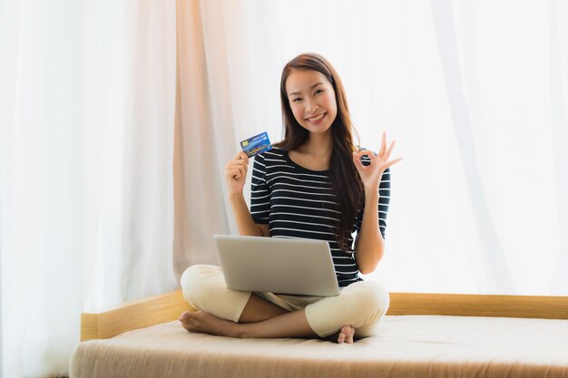 Mujer asiática joven hermosa del retrato que usa el cuaderno o el ordenador portátil del ordenador con la tarjeta de crédito para hacer compras