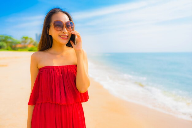 Mujer asiática joven hermosa del retrato que usa el teléfono móvil o el teléfono móvil en la playa y el mar