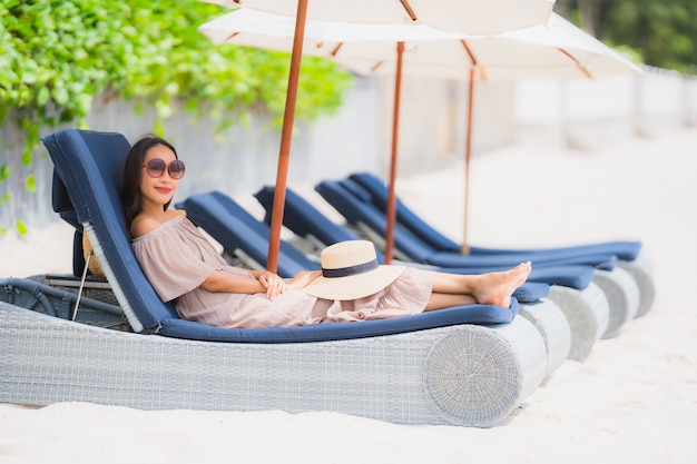 Mujer asiática joven hermosa del retrato en la silla de cubierta con el paraguas alrededor del océano del mar de la playa