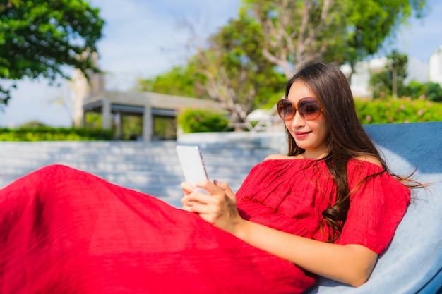 Mujer asiática joven hermosa del retrato con el teléfono celular o el teléfono móvil alrededor de piscina en el centro turístico del hotel