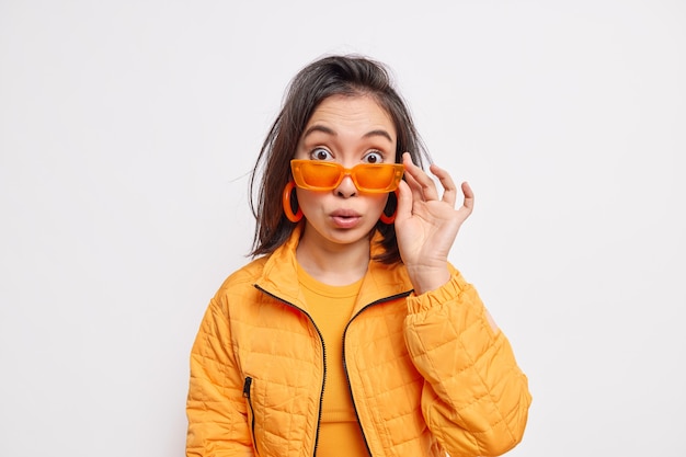 La mujer asiática joven morena de moda sorprendida lleva la chaqueta anaranjada de las gafas de sol de moda y los pendientes reacciona en algo asombroso aislado sobre la pared blanca. Concepto de estilo y moda.