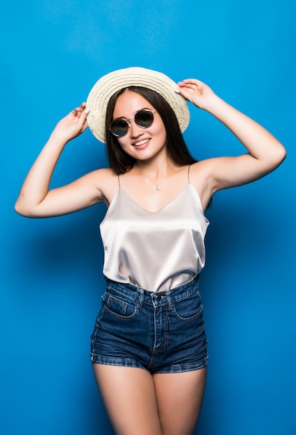 Mujer asiática joven con pose de sorpresa aislado sobre fondo azul. Retrato de hermosa mujer asiática en sombrero de paja y gafas de sol sobre fondo azul.