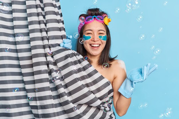 Mujer asiática joven positiva aplica parches de colágeno de hidrogel debajo de los ojos rizadores de pelo posa detrás de la cortina de la ducha disfruta de los procedimientos de higiene