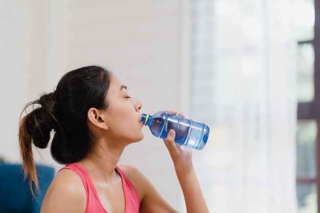 Mujer asiática joven que bebe agua porque siente descanso agotado después de hacer ejercicio en la sala de estar