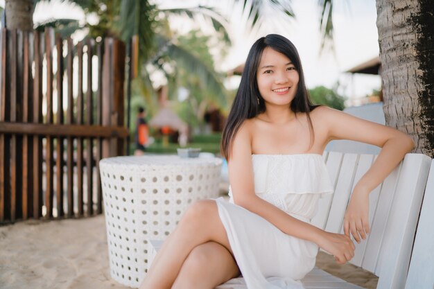 La mujer asiática joven que se sienta en banco se relaja en la playa, feliz femenino hermoso se relaja cerca del mar.