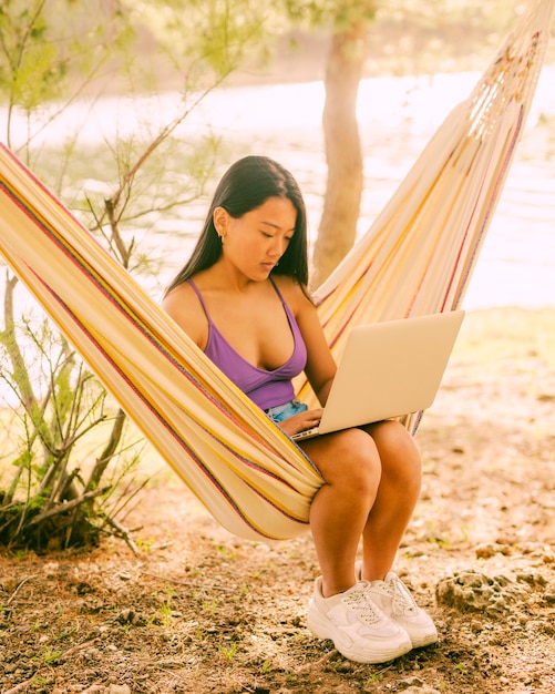 Mujer asiática joven que se sienta en hamaca cerca del río y que trabaja independientemente en la computadora portátil