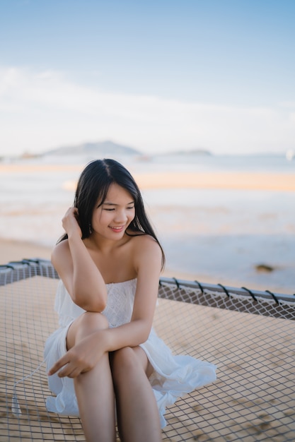 La mujer asiática joven que se sienta en la hamaca se relaja en la playa, feliz femenino hermoso se relaja cerca del mar.