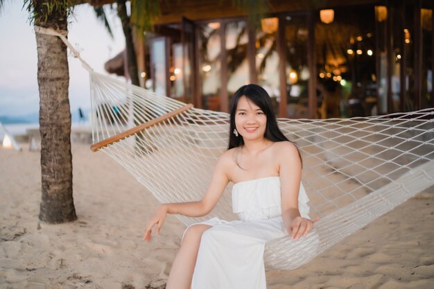 La mujer asiática joven que se sienta en la hamaca se relaja en la playa, feliz femenino hermoso se relaja cerca del mar.