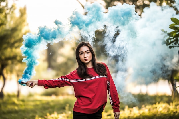 Mujer asiática joven que sostiene la bomba de humo colorida azul en el parque al aire libre. Propagación de humo azul