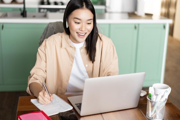 Mujer asiática joven que toma notas trabajando durante la videoconferencia con la computadora portátil en casa