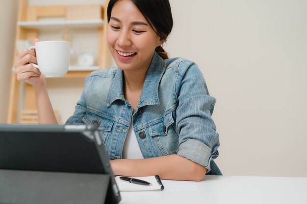 Mujer asiática joven que trabaja usando la tableta que comprueba medios sociales y que bebe el café mientras que relájese en el escritorio en sala de estar en casa.