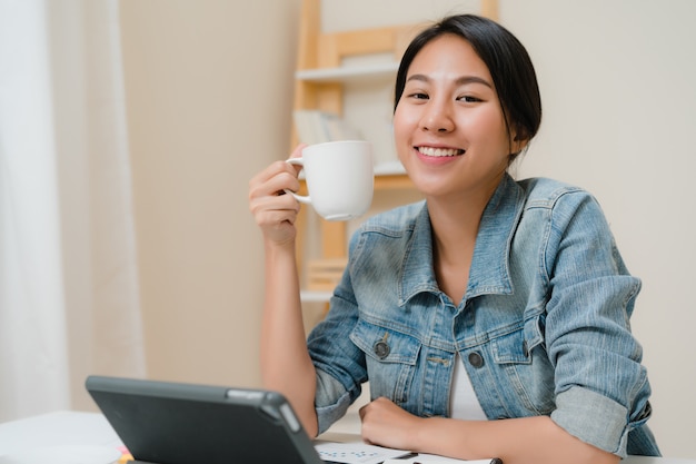 Mujer asiática joven que trabaja usando la tableta que comprueba medios sociales y que bebe el café mientras que relájese en el escritorio en sala de estar en casa.
