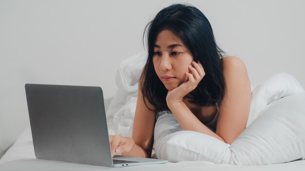 La mujer asiática joven que usa la computadora portátil que comprueba medios sociales que siente feliz sonriendo mientras que miente en cama después de despertar en la casa en la mañana, muchacha tailandesa atractiva que sonríe se relaja en dormitorio en casa.