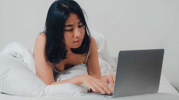 La mujer asiática joven que usa la computadora portátil que comprueba medios sociales que siente feliz sonriendo mientras que miente en cama después de despertar en la casa en la mañana, muchacha tailandesa atractiva que sonríe se relaja en dormitorio en casa.