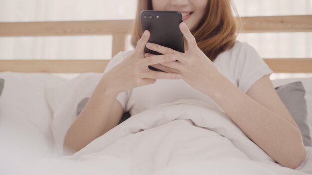 La mujer asiática joven que usa smartphone mientras que miente en cama después de despierta por la mañana