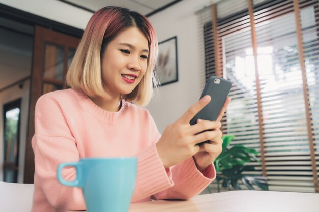 Mujer asiática joven que usa smartphone mientras que miente en el escritorio en su sala de estar