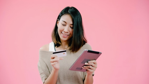 Mujer asiática joven que usa la tableta que compra compras en línea por la tarjeta de crédito que siente la sonrisa feliz en ropa informal sobre tiro rosado del estudio del fondo. La mujer alegre adorable sonriente feliz disfruta de éxito