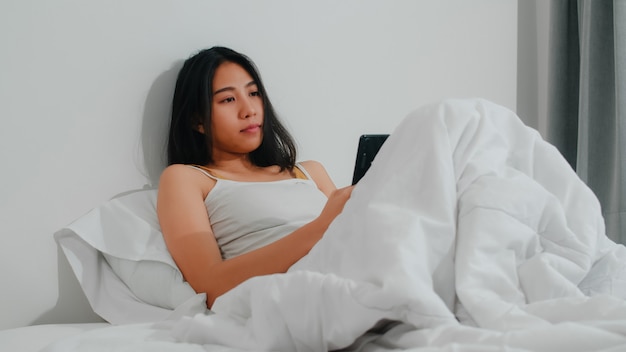 Mujer asiática joven que usa el teléfono inteligente que comprueba los medios sociales que se siente feliz sonriendo mientras está acostado en la cama después de despertarse por la mañana, bella dama hispana atractiva sonriendo relajarse en el dormitorio en casa.
