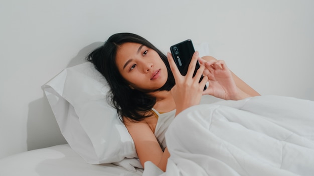 Mujer asiática joven que usa el teléfono inteligente que comprueba los medios sociales que se siente feliz sonriendo mientras está acostado en la cama después de despertarse por la mañana, bella dama hispana atractiva sonriendo relajarse en el dormitorio en casa.