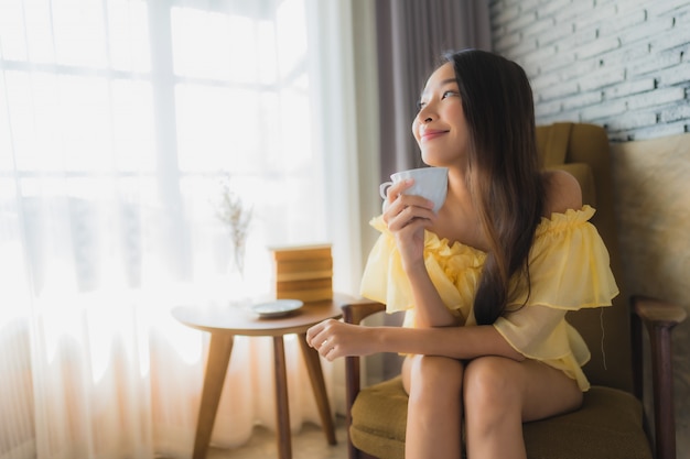 La mujer asiática joven del retrato se sienta en la silla del sofá y lee el libro con la taza de café