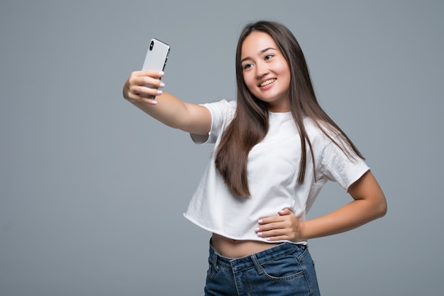 Mujer asiática joven sonriente que toma un selfie con el teléfono móvil sobre fondo gris aislado de la pared